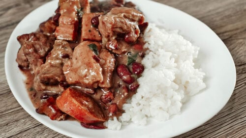 Hearty bean stew with pork and smoked sausages served over white rice on a white plate.