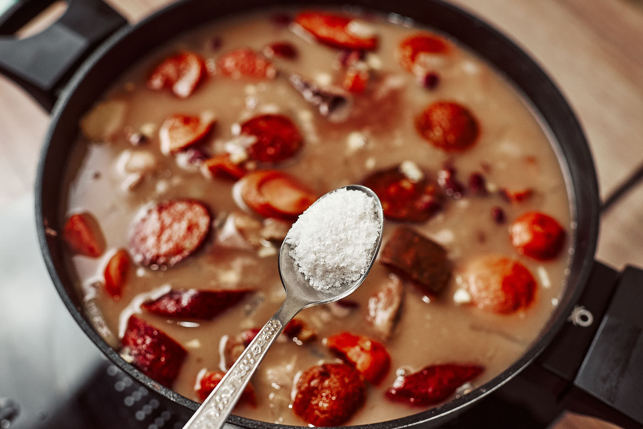 Adding a spoonful of salt to a pot of simmering bean stew with pork and smoked sausages.