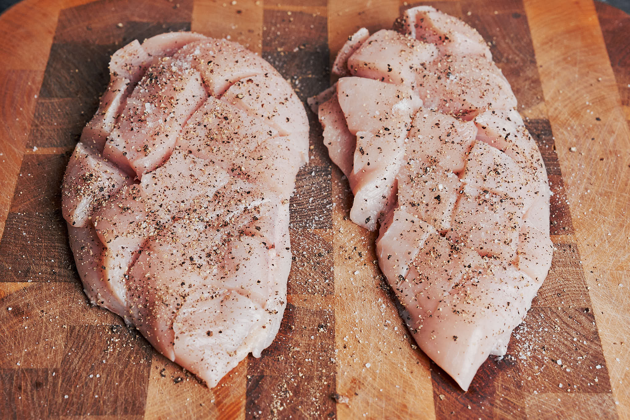 Raw chicken with crosshatch cuts seasoned with salt and pepper on a wooden cutting board