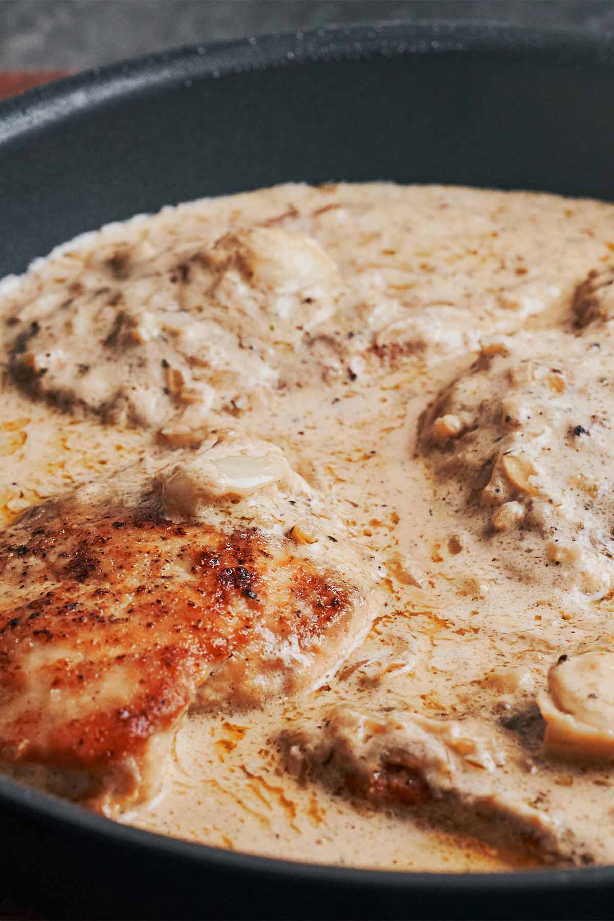 Close-up of creamy garlic chicken breasts in a skillet with whole garlic cloves and a rich, silky cream sauce