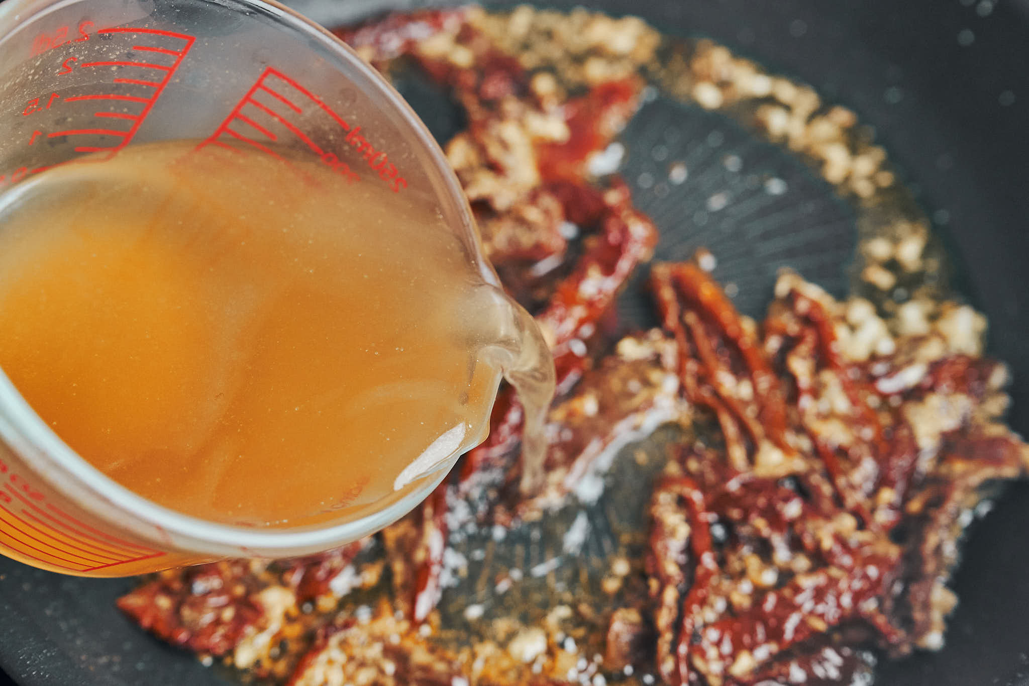 Chicken broth being poured into the skillet with sun-dried tomatoes