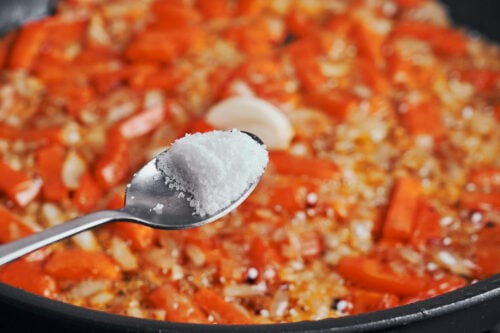 a spoonful of salt being added to the skillet vegetables