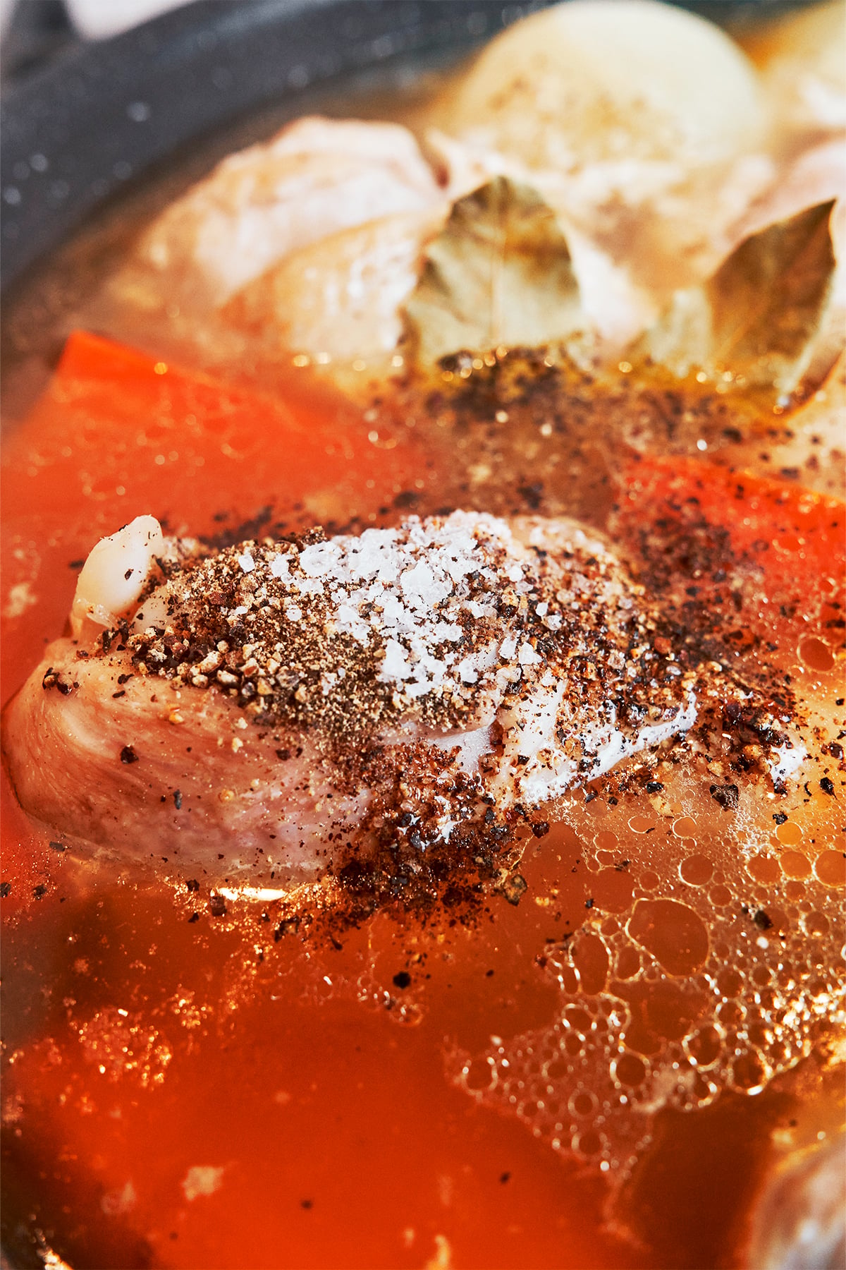 Chicken simmering in warm homemade broth with carrots, bay leaves, salt, and black pepper