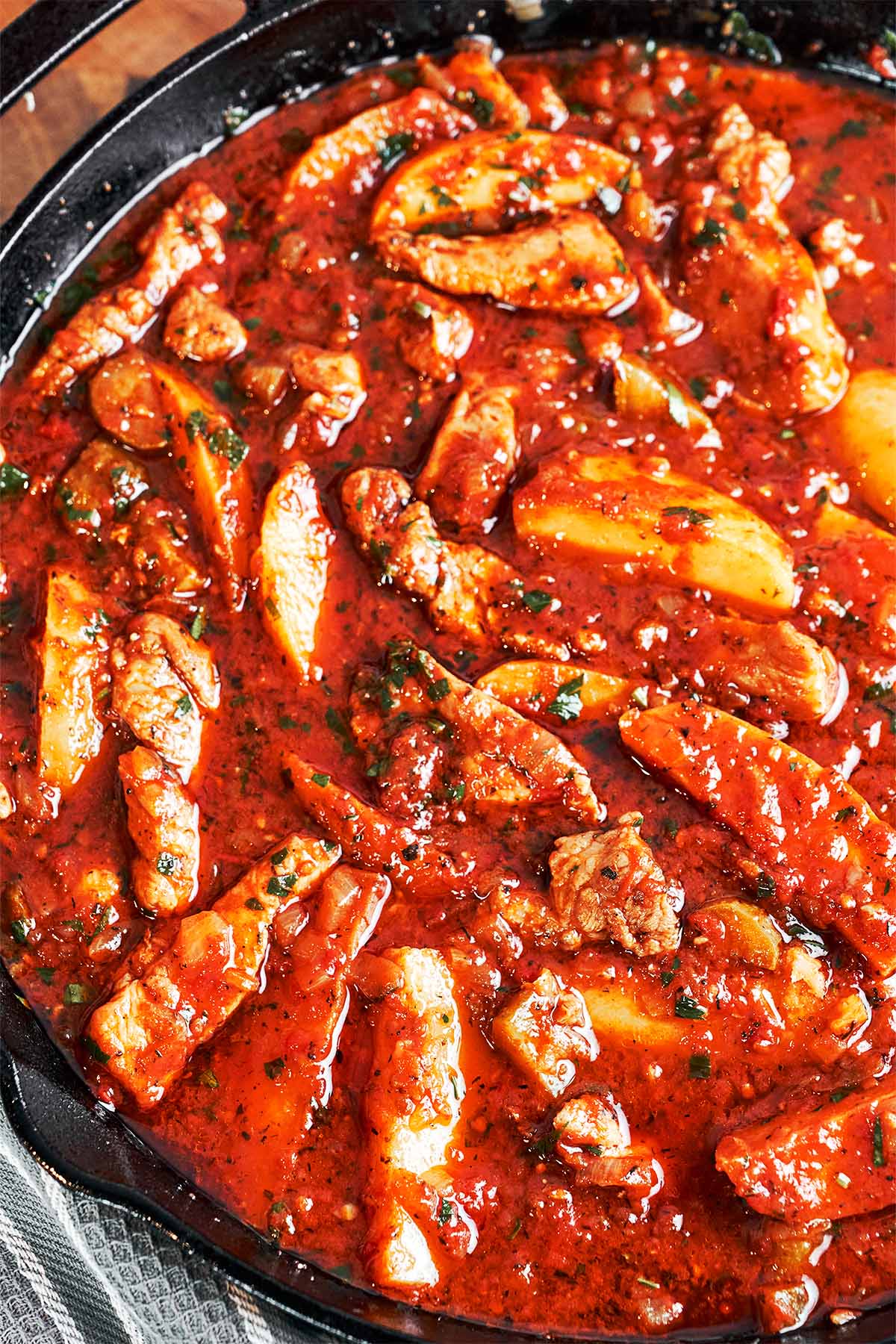 Close-up of tender pork and golden potato wedges coated in a glossy tomato-pickle sauce with fresh parsley.