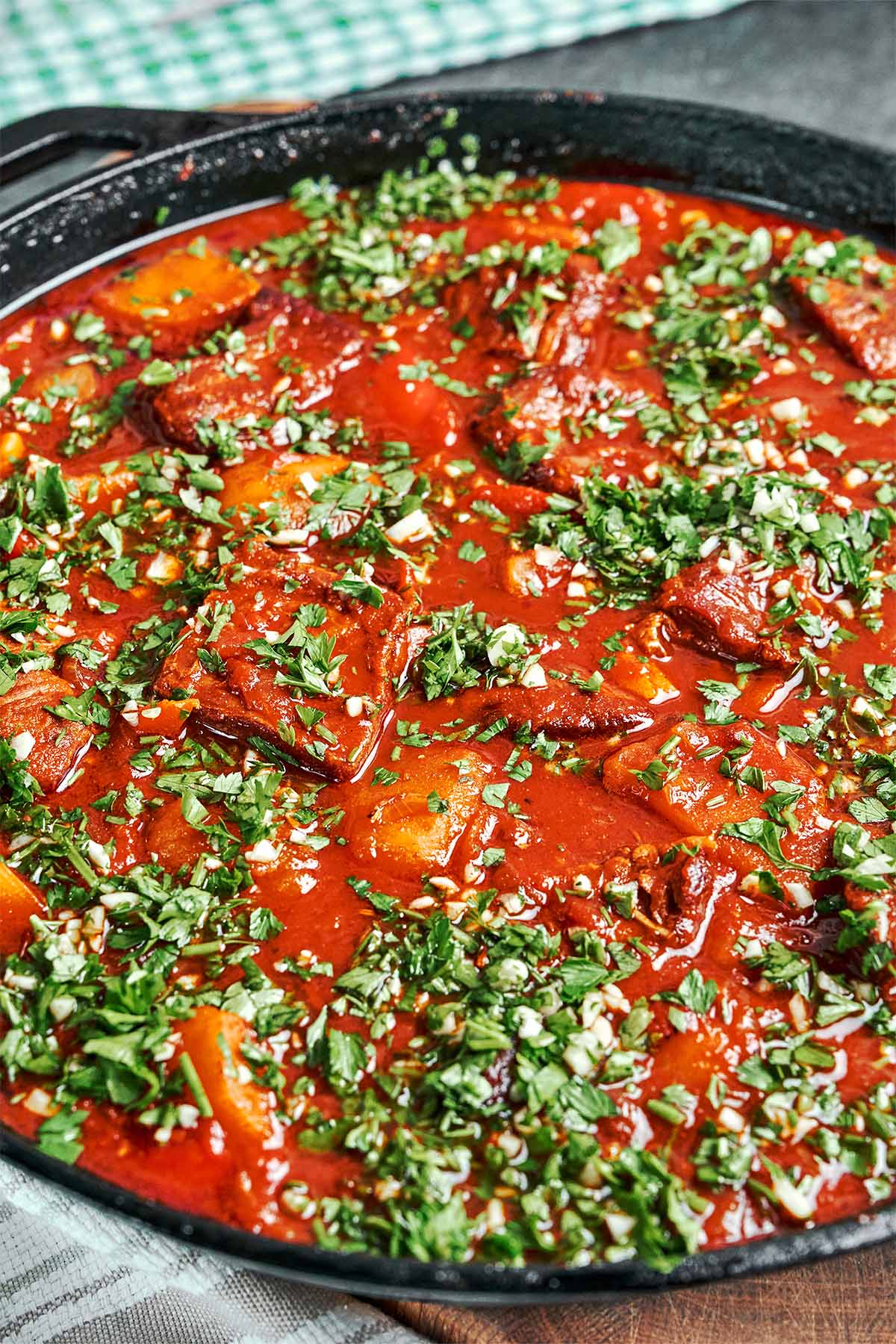Beef stew with vegetables in a rich tomato sauce, topped with freshly chopped parsley and garlic in a black skillet.
