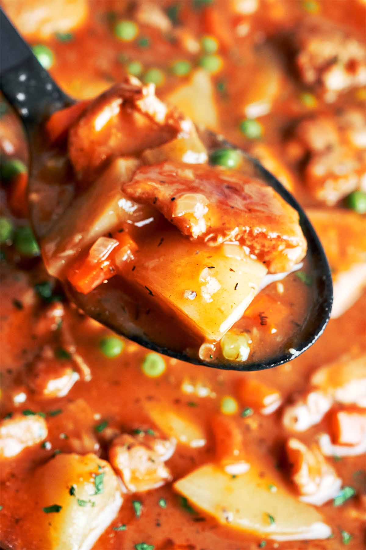 A spoonful of tender chicken and potato lifted from the rich, savory stew with carrots and peas in the background.
