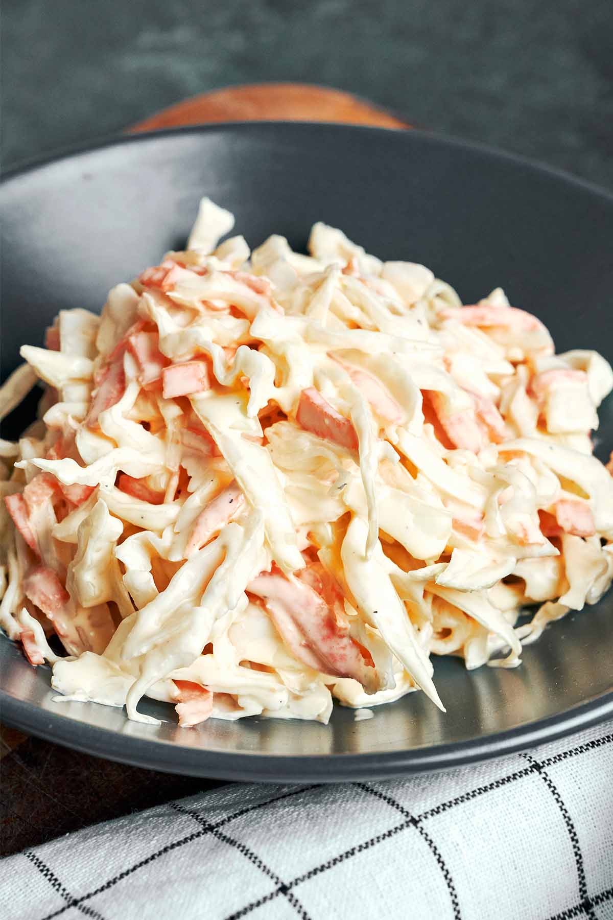 Creamy coleslaw piled onto a dark plate, with ribbons of cabbage and orange carrots coated in a smooth sour cream dressing.