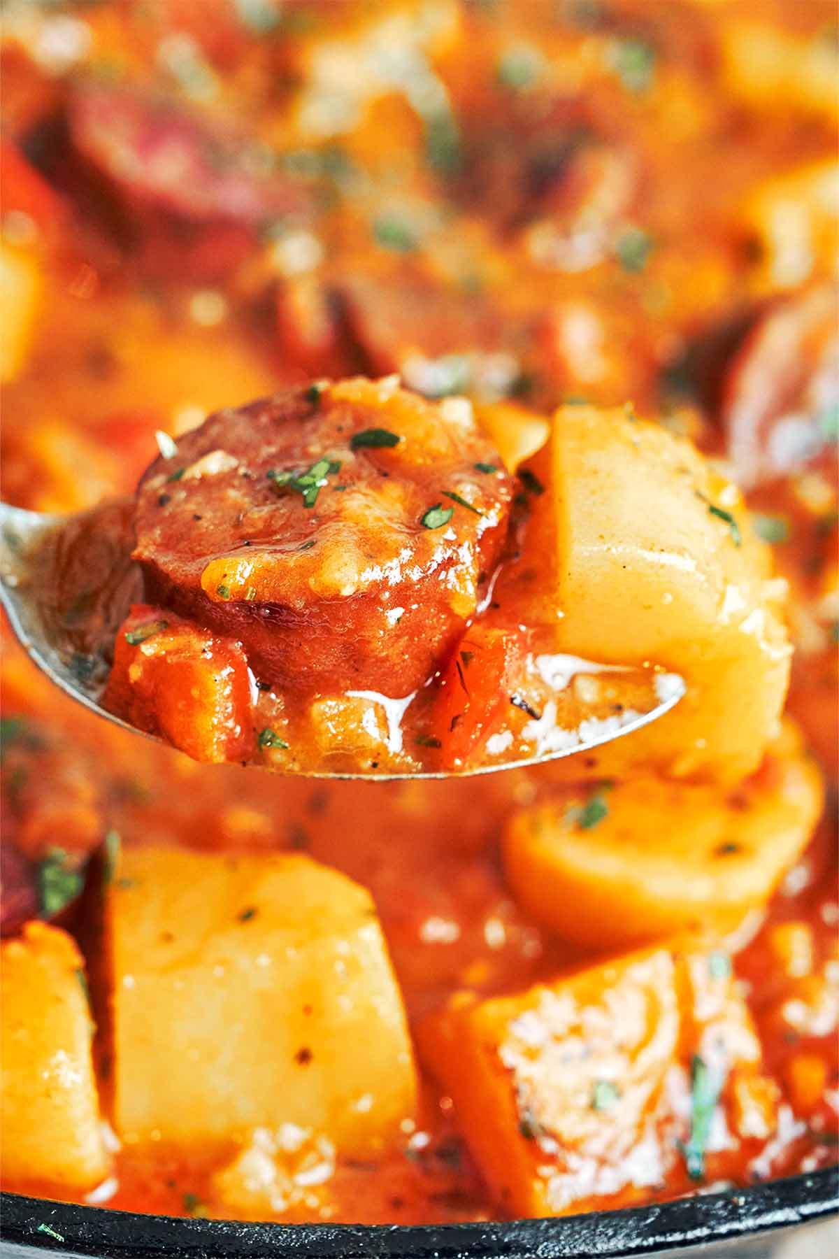 A spoonful of smoked sausage, tender potato, and sweet vegetables coated in paprika sauce, lifted from the skillet.